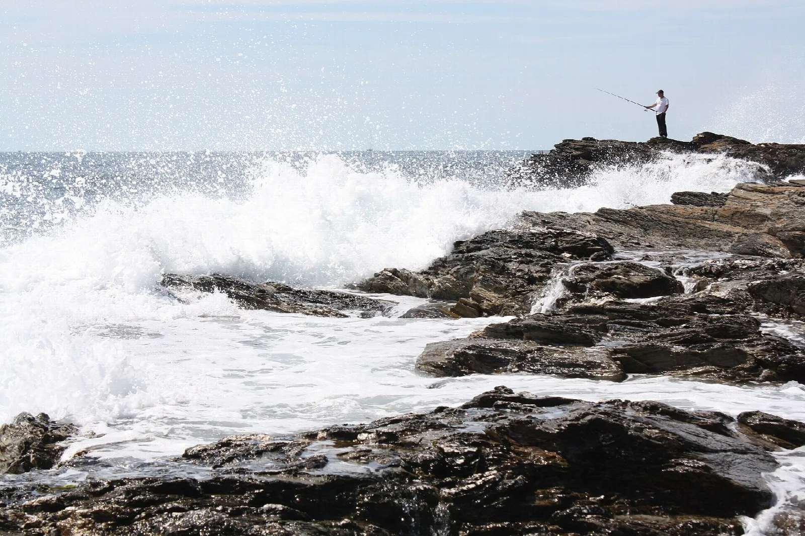 Waves crashing over dark rocks with a person fishing at the edge
