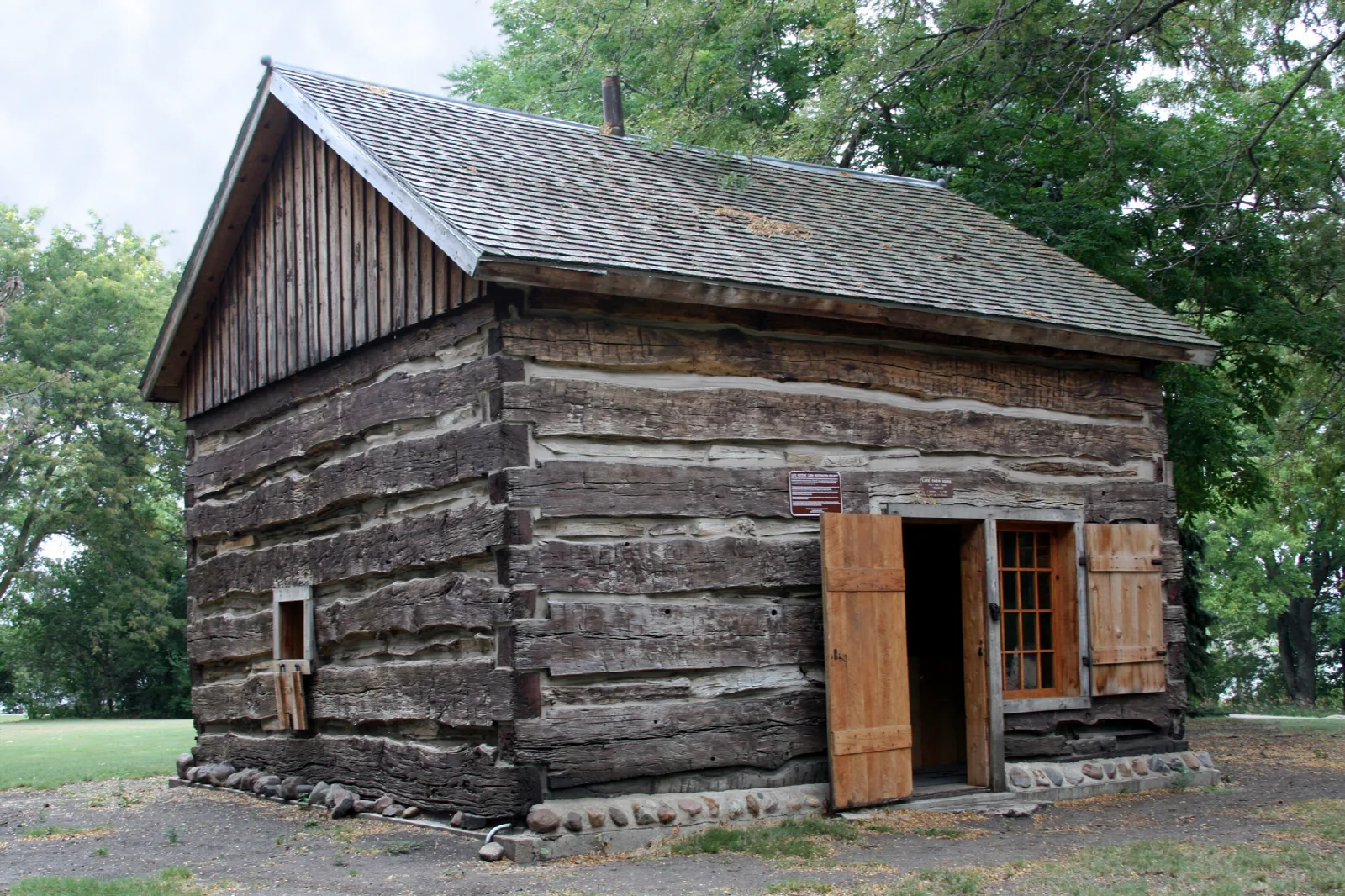 Old log cabin with a shingled roof and open wooden shutters among trees
