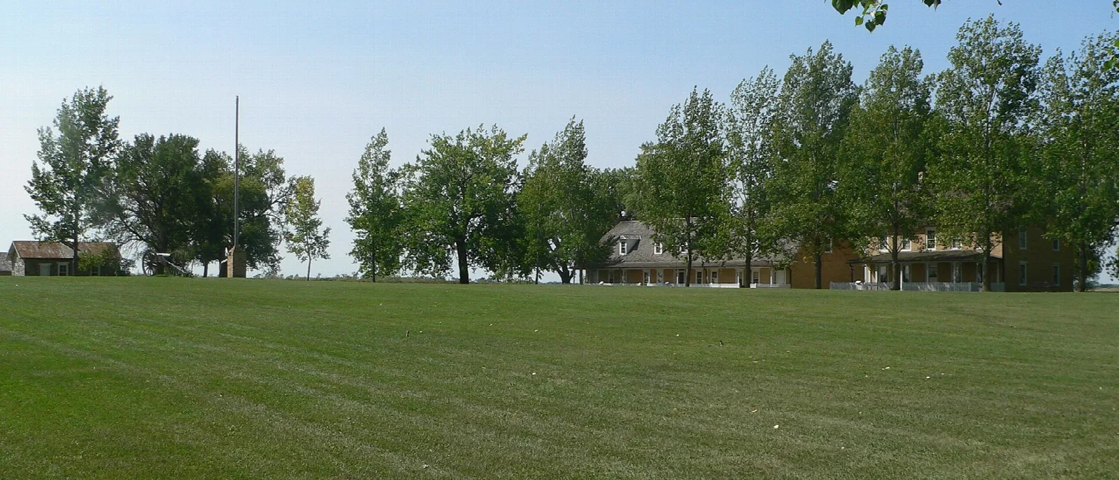 Wide green parade ground with fort buildings among trees beyond