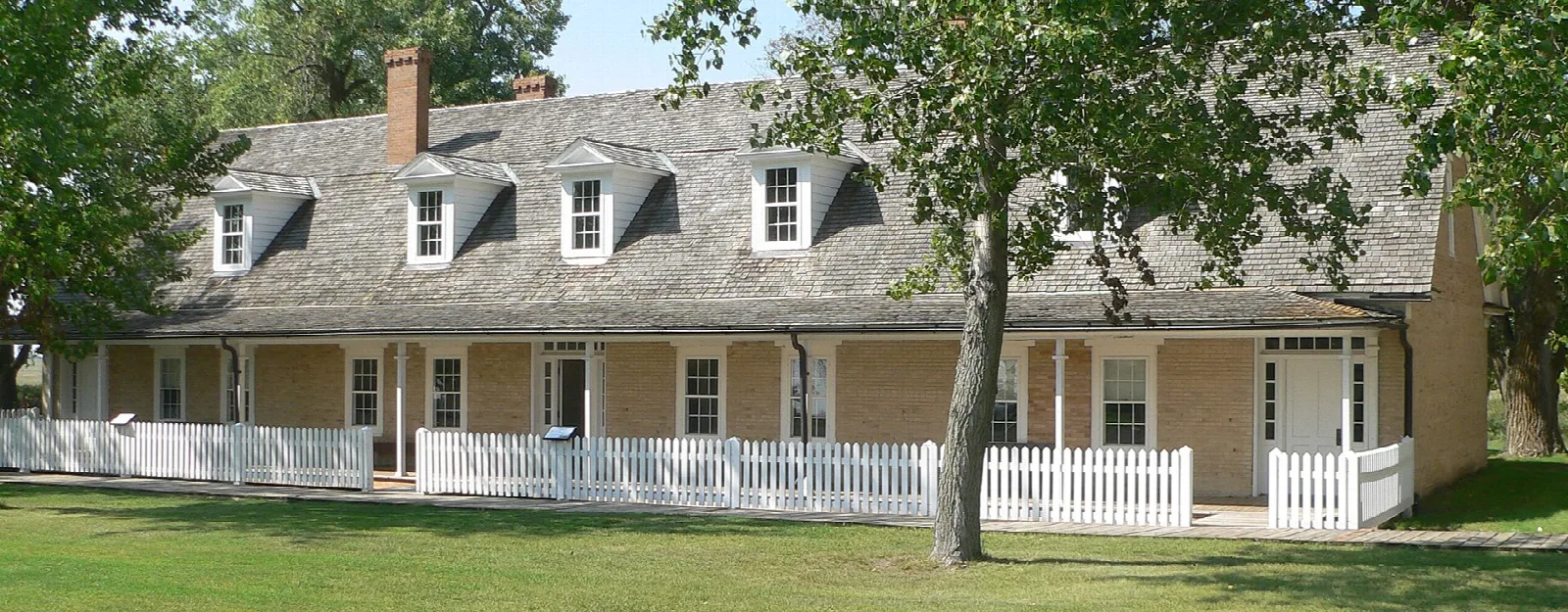 Long officers' quarters with dormer windows, a porch and a white fence under a tree