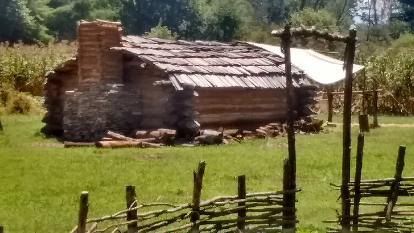 Log cabin with a stone chimney behind a woven fence in a grassy field