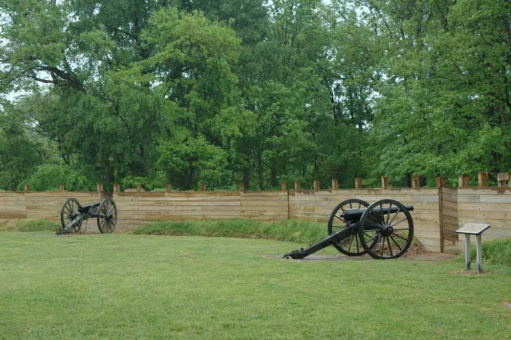 Two cannons on grass in front of a wooden fort wall and trees