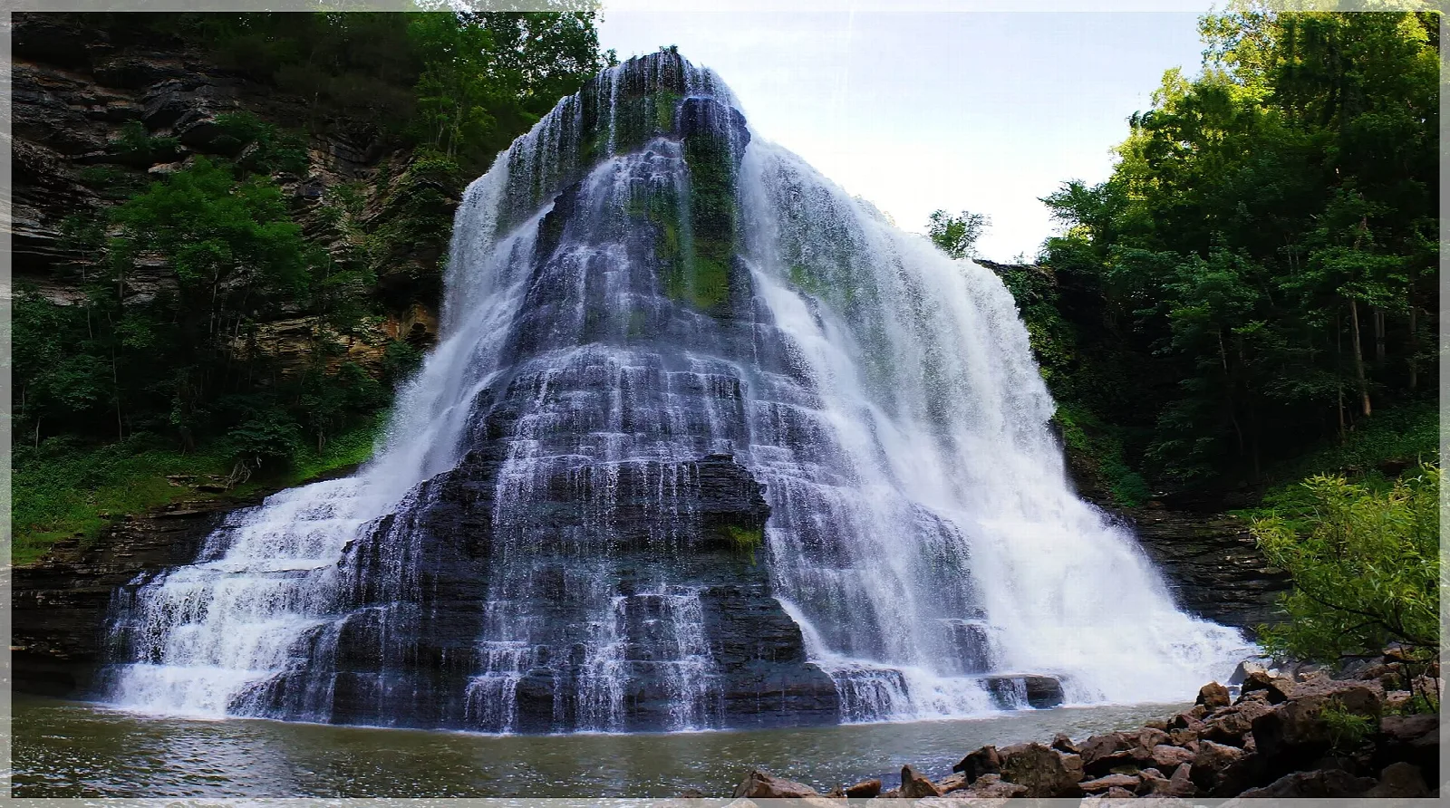 Tiered waterfall spreading down dark rock into a pool with green trees