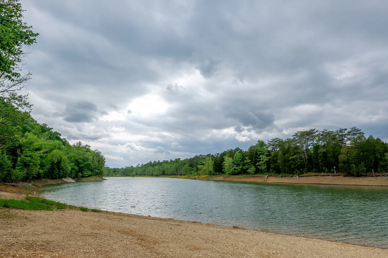 Wide lake under cloudy sky with a sandy shore and forested hills