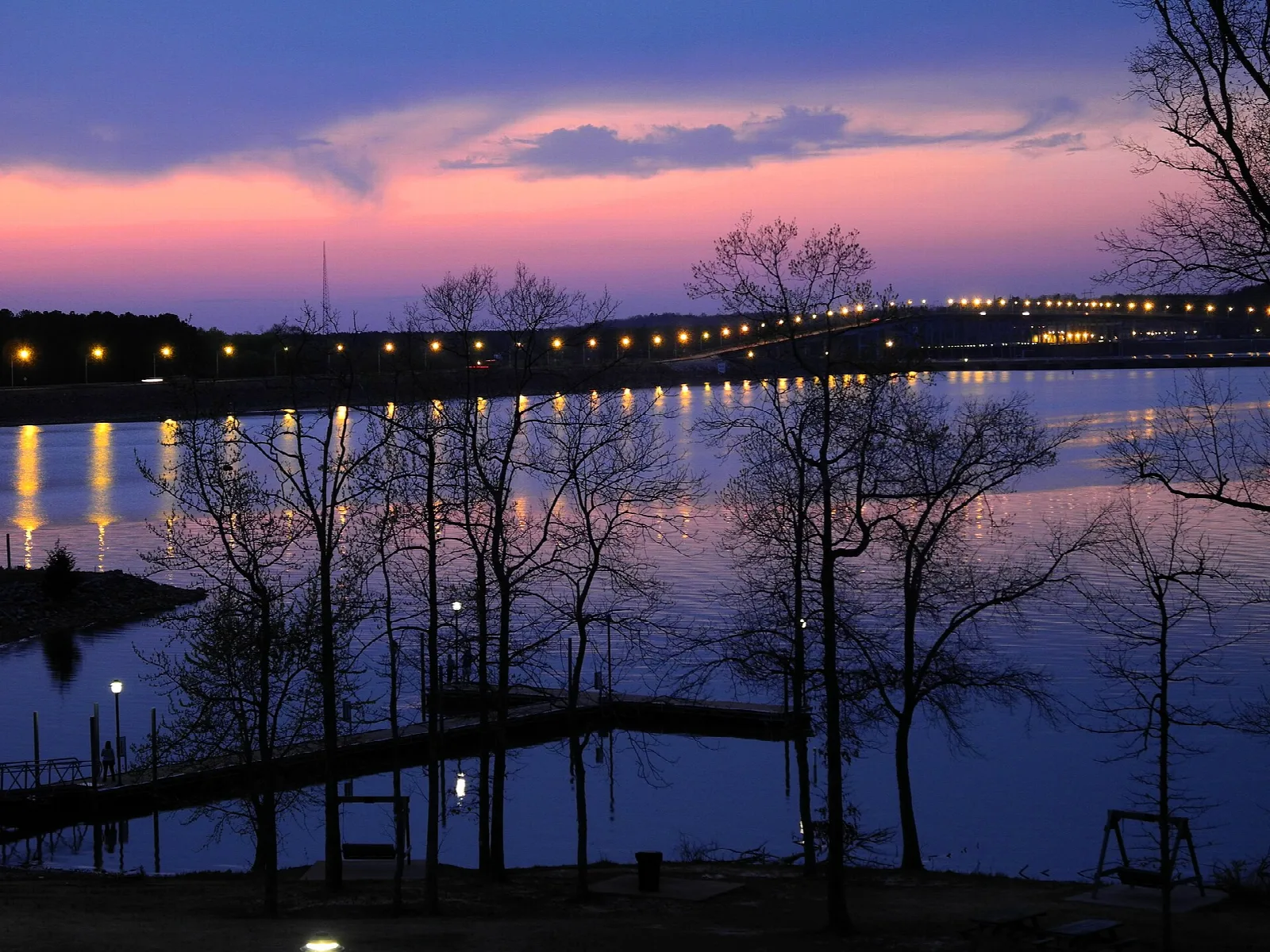 Dusk sky over a lake with a lit dam in the distance and bare trees
