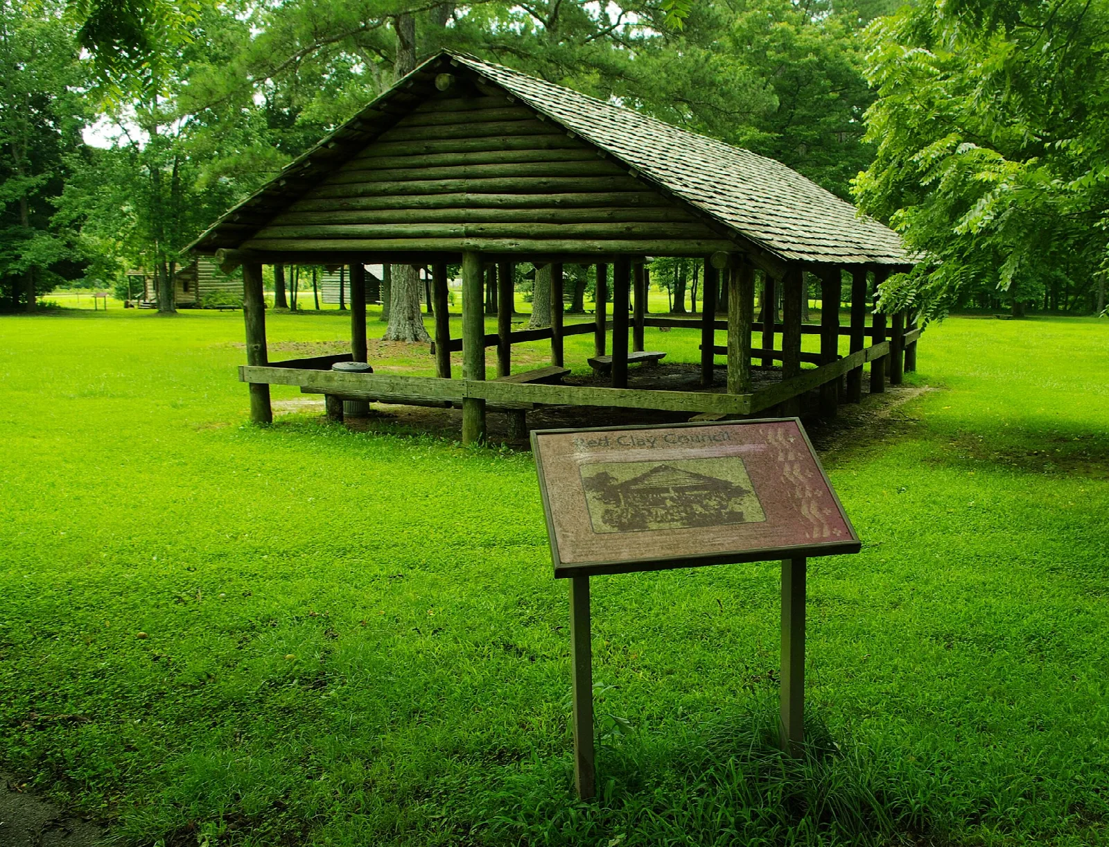 Open-sided log council house with a roof on a green lawn, beside an interpretive sign