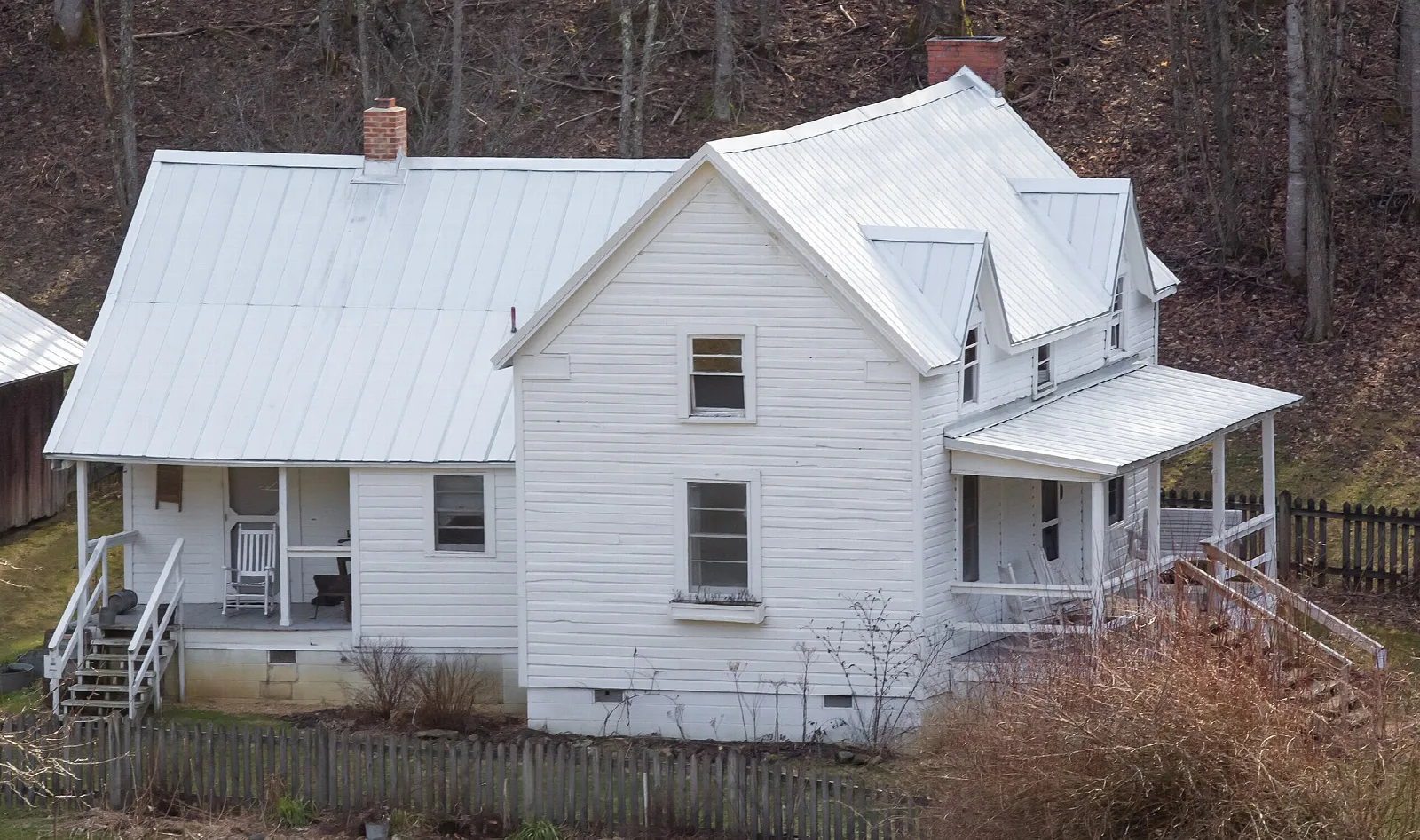 White farmhouse with a metal roof and porch on a wooded hillside