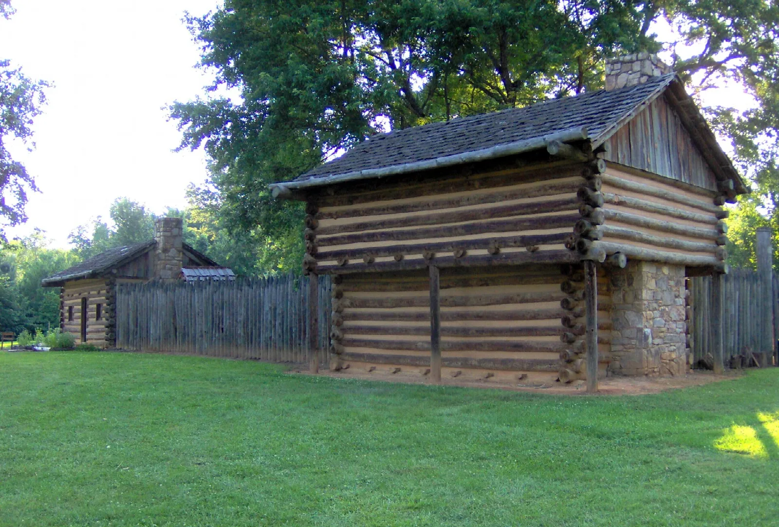 Log blockhouse with a stone base inside a wooden palisade on a green lawn