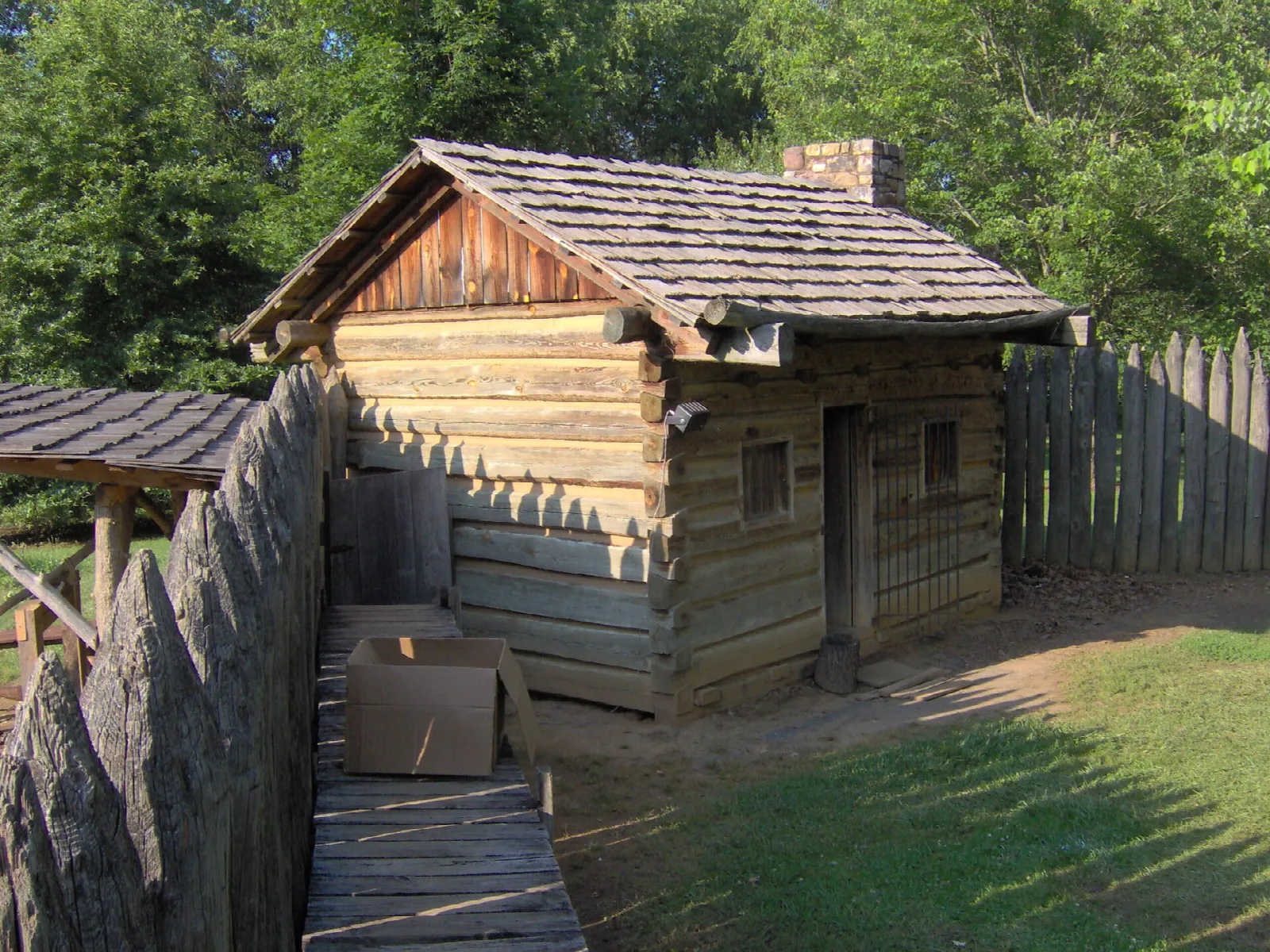 Log cabin with a shingled roof behind a wooden palisade