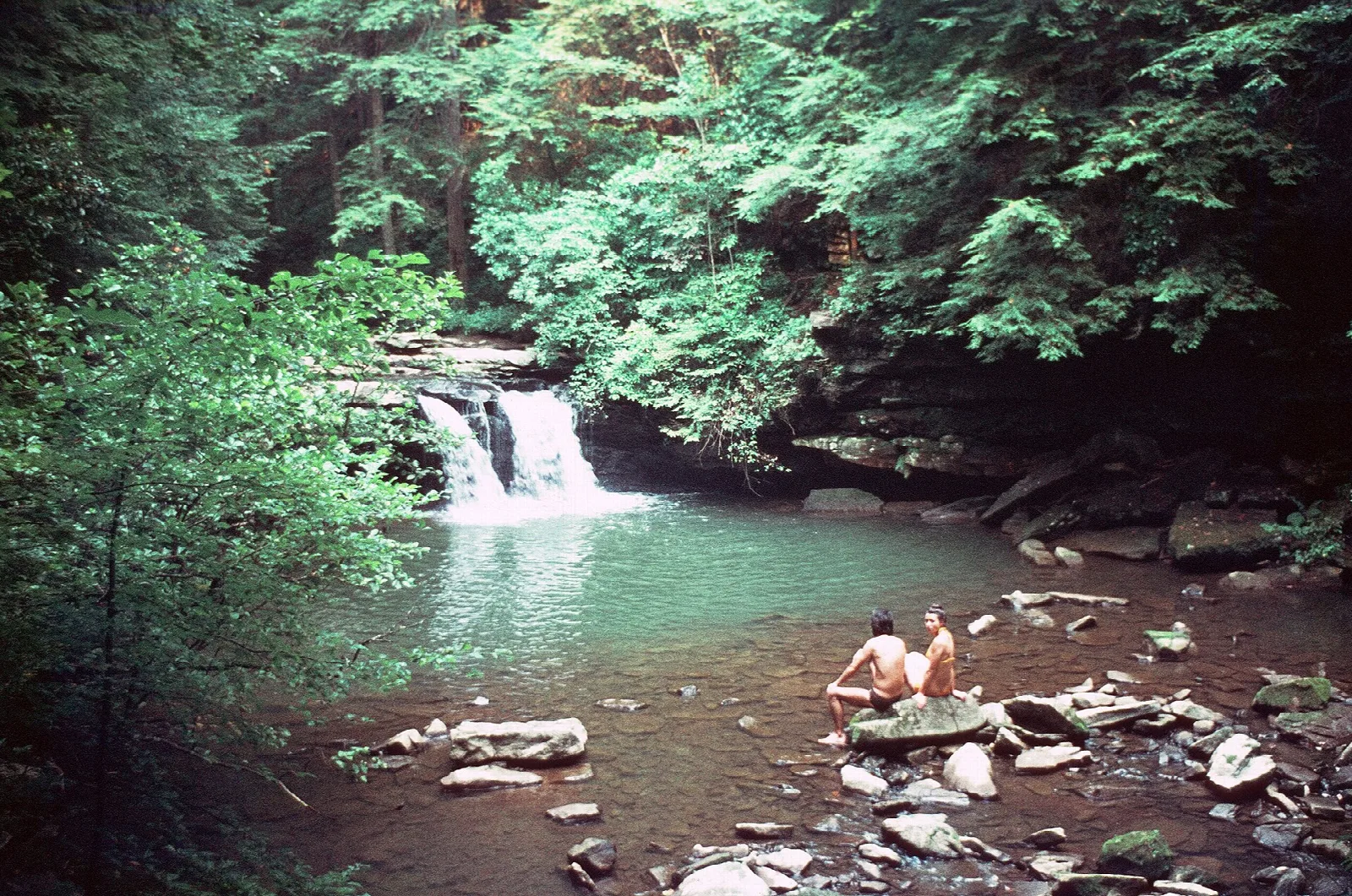Small waterfall and green pool below a rock ledge, with two people sitting