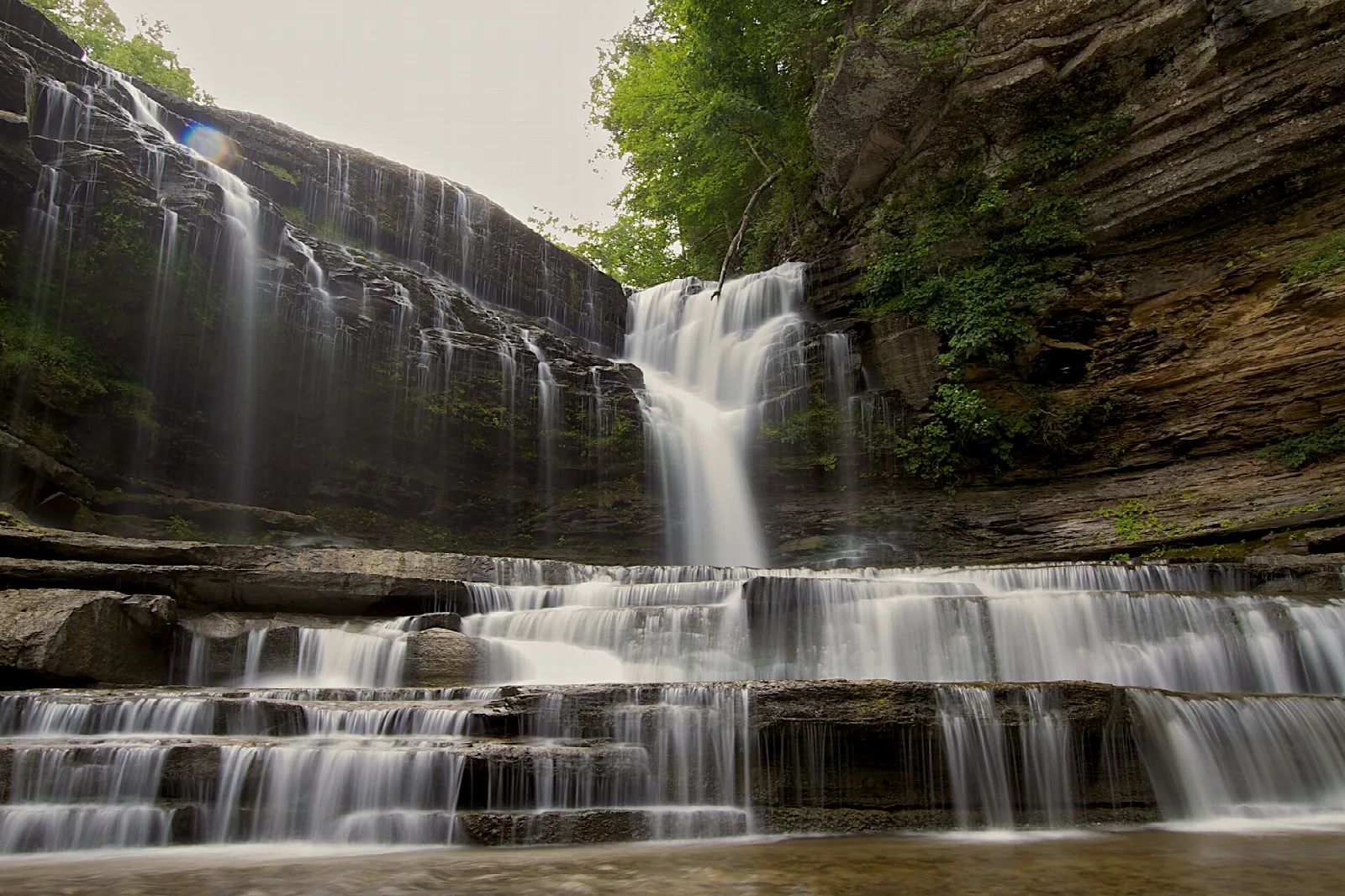 Wide tiered waterfall pouring over rock ledges in a wooded gorge