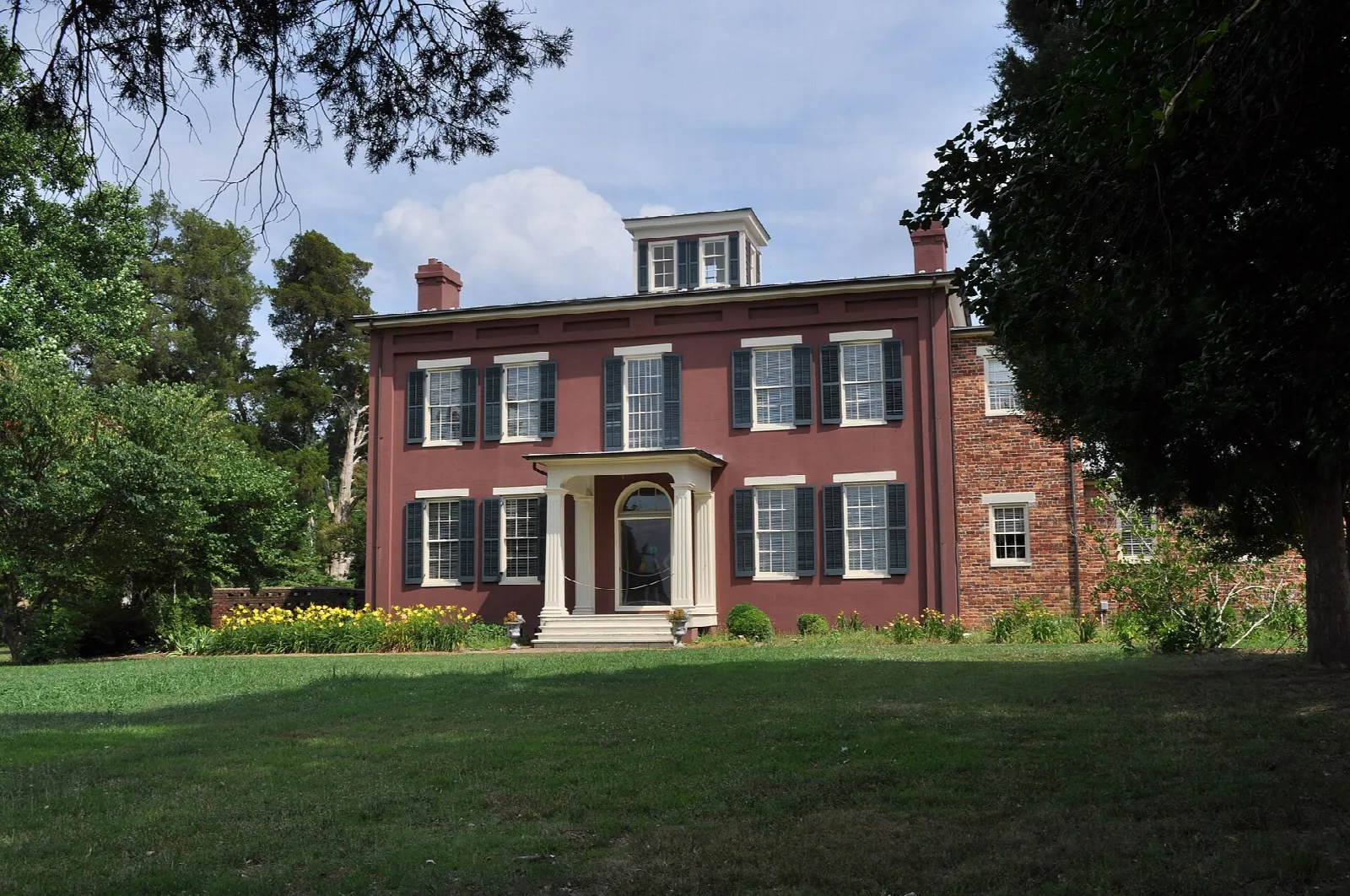 Red brick mansion with white trim behind a wide green lawn and large trees