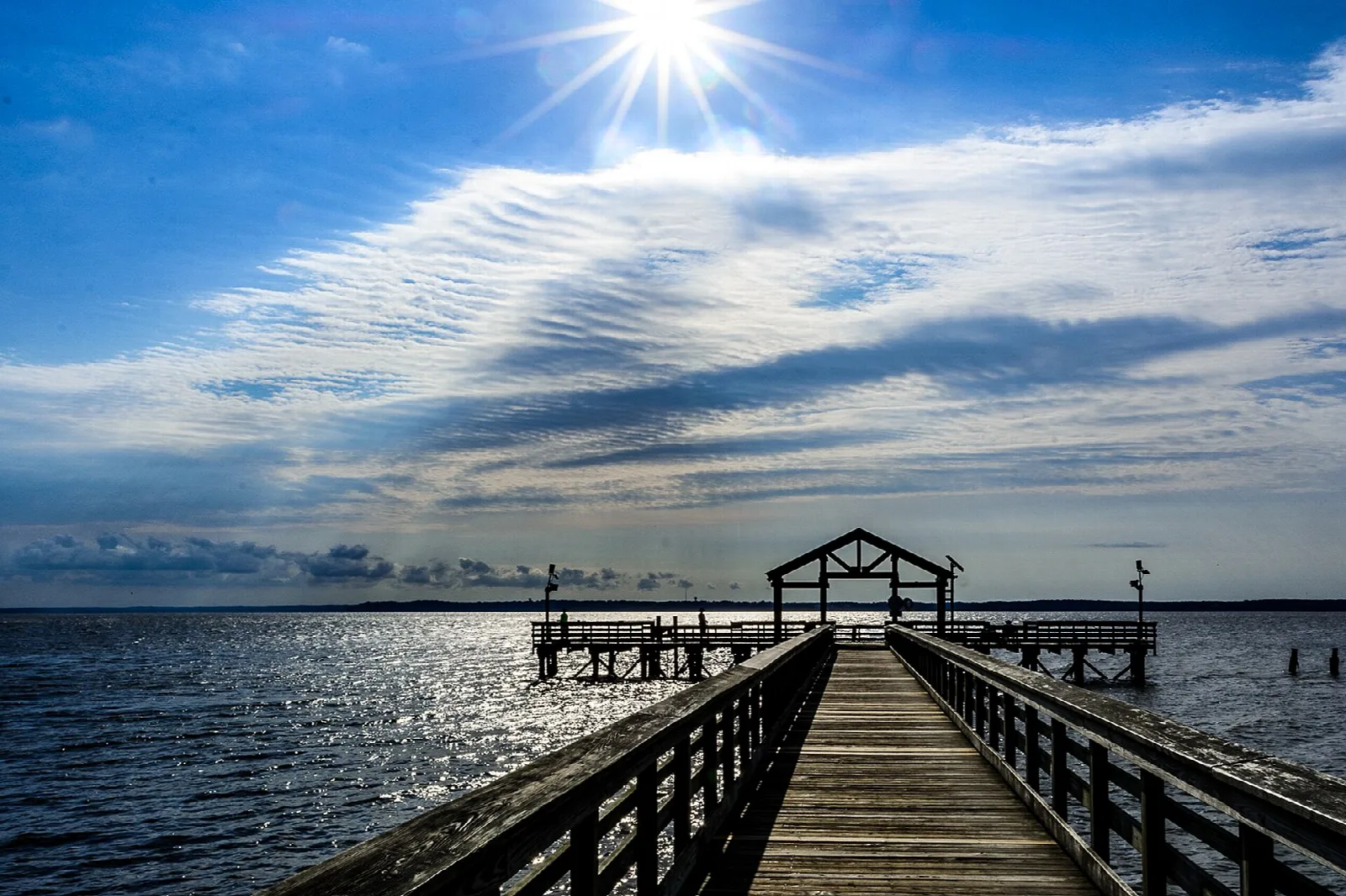 Long wooden fishing pier over the river under a bright sunburst sky