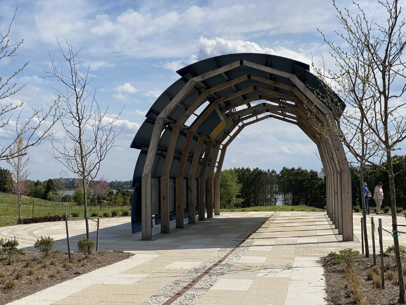 Arched wooden pavilion on a paved path under a partly cloudy sky