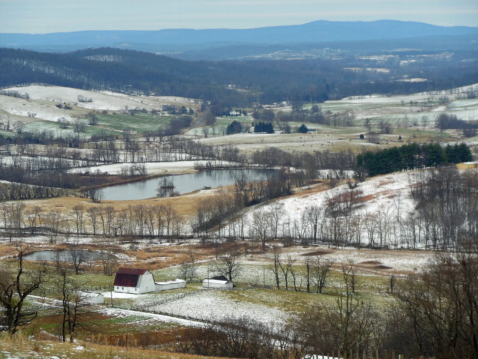 Snow-dusted farm fields and a pond in a valley below blue ridges