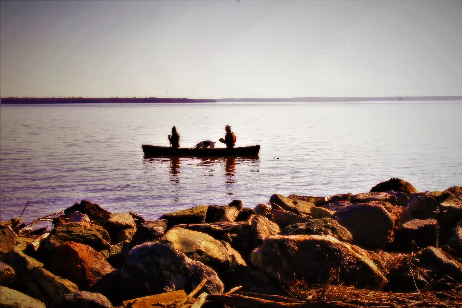Two people in a canoe on calm water at dusk beyond a rocky shore