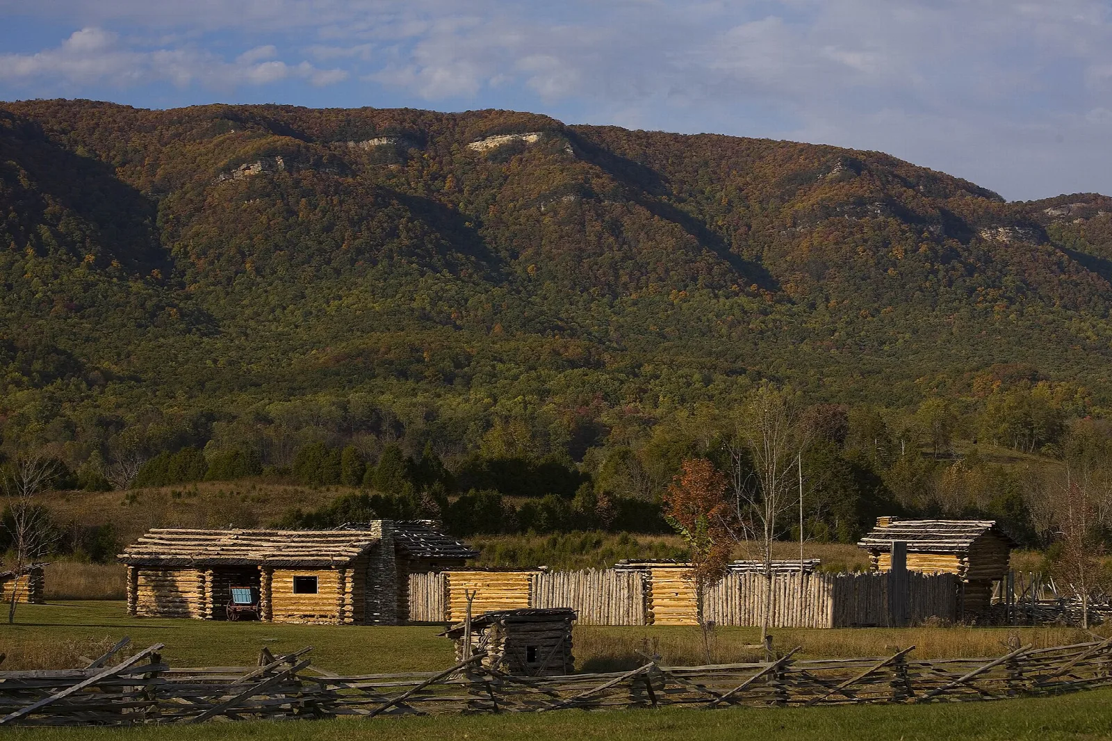 Log buildings of a frontier fort below a long wooded mountain ridge