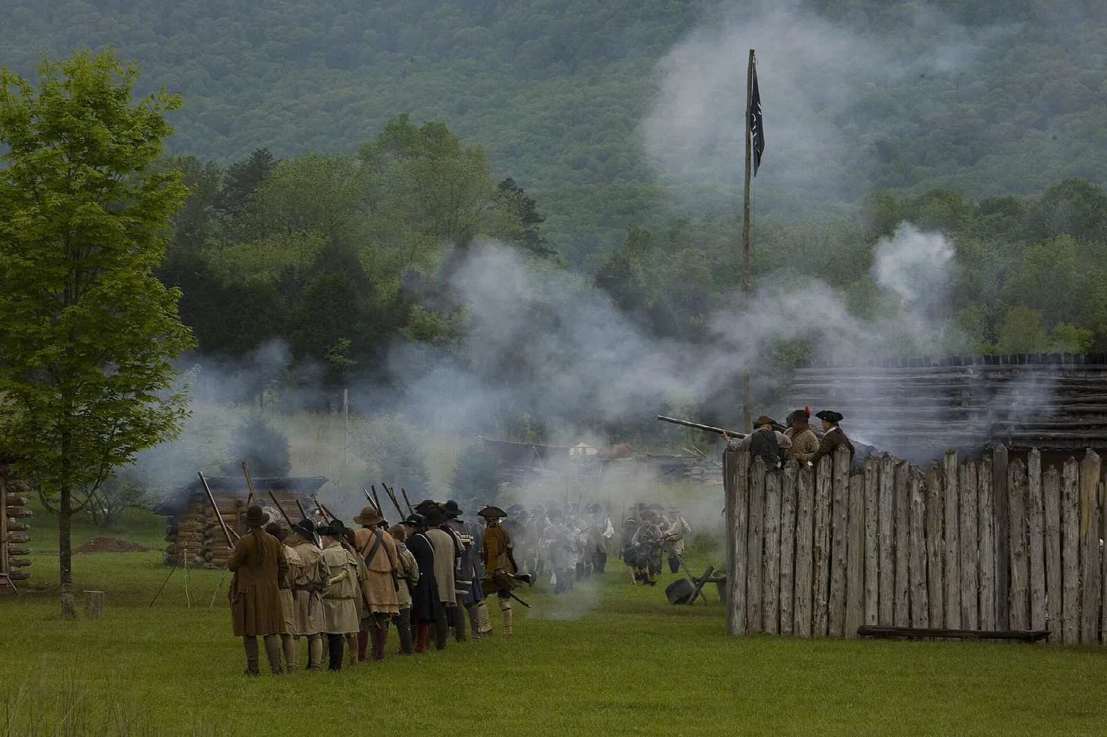 Smoke rising over a log stockade as costumed reenactors stand nearby