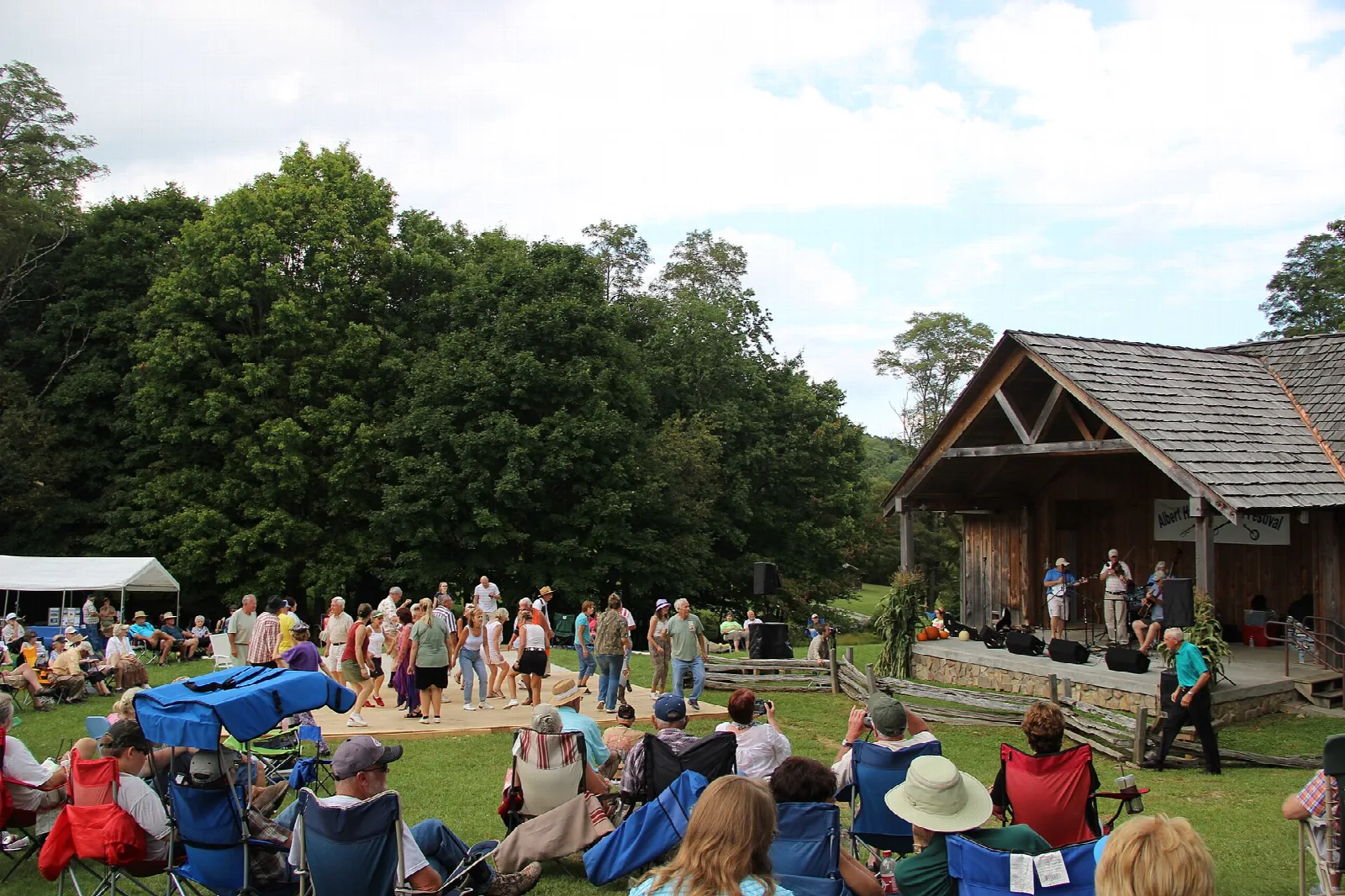 Crowd seated on a lawn watching a stage beside a log building