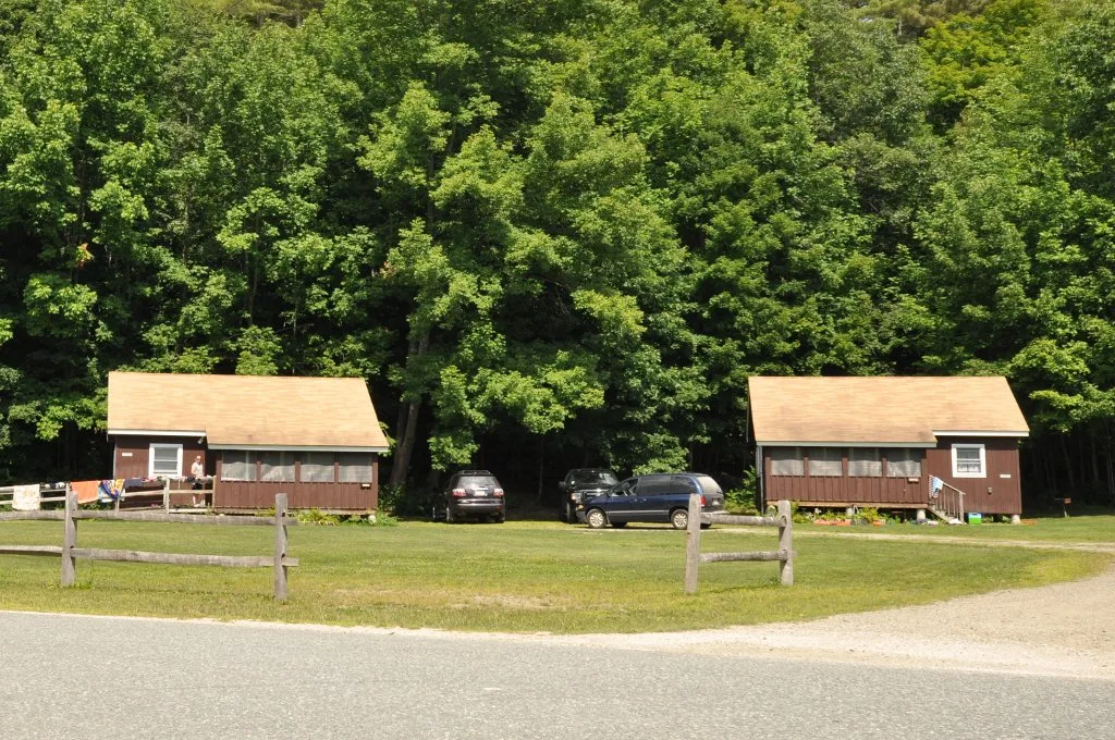 Two small brown cabins with shingle roofs on a lawn in front of dense green trees