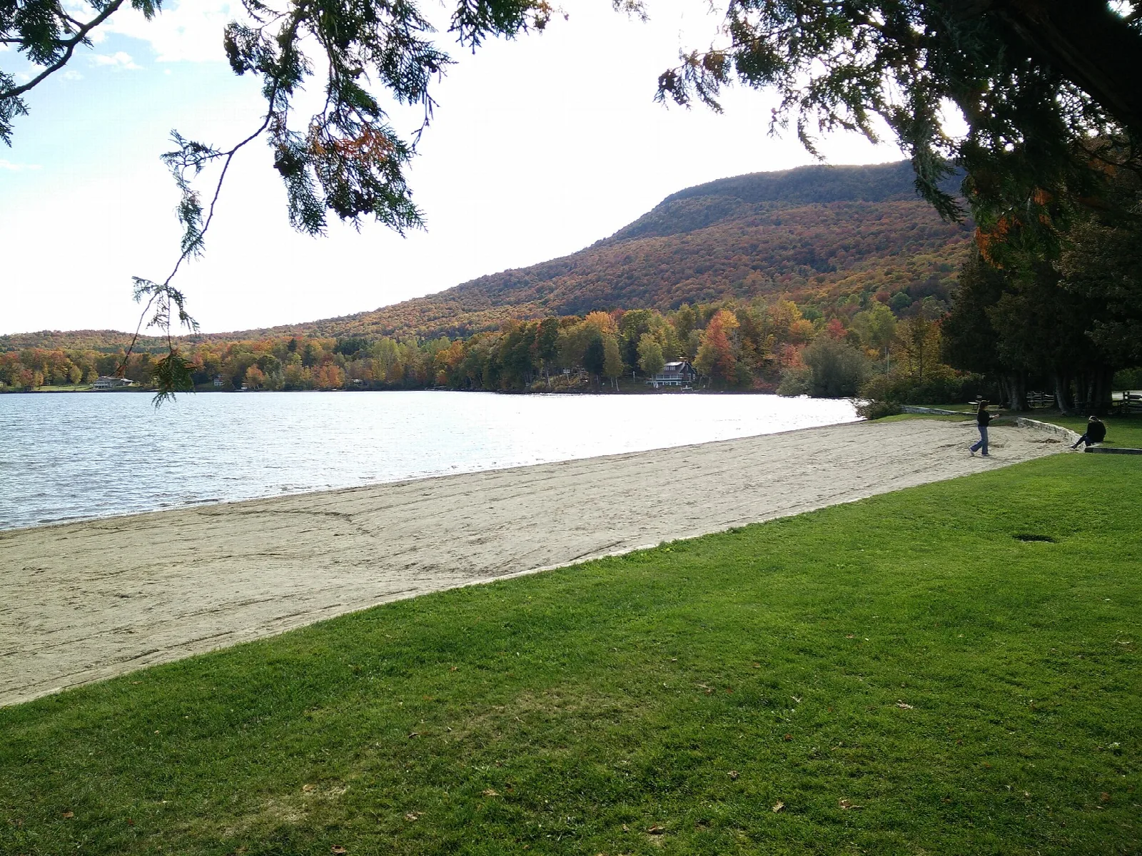 Sandy beach and grass lawn along a lake, with a wooded hill in autumn color beyond