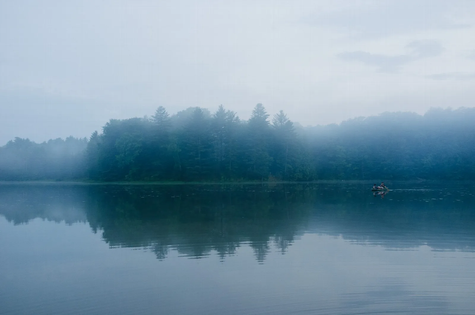 Misty pond reflecting a line of evergreen trees, with one small boat on the water
