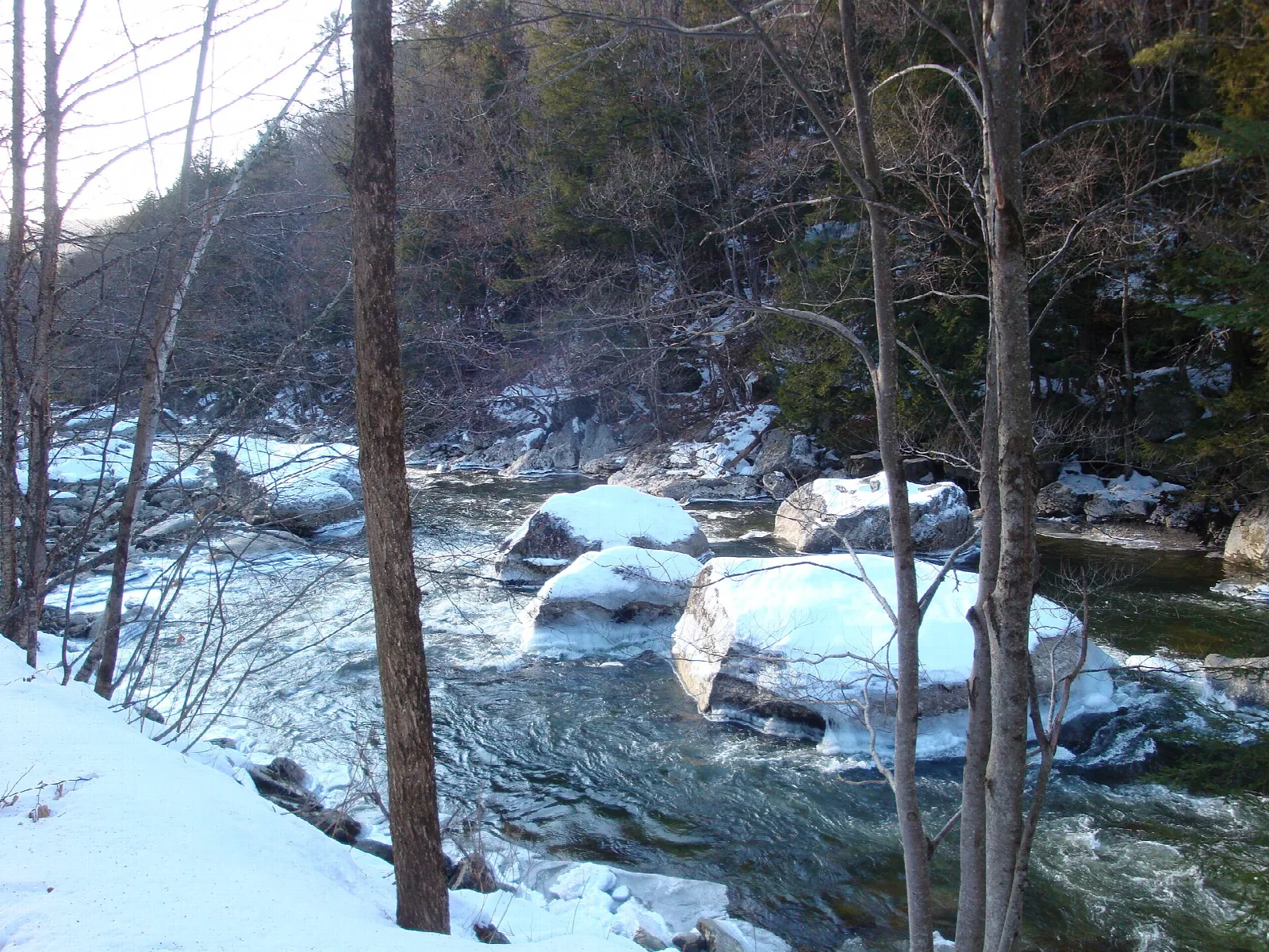 Snow-covered boulders in a rushing river below bare trees