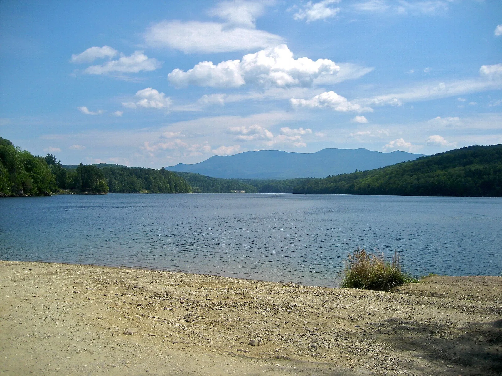 Sandy lake shore with blue water and forested mountains in the distance