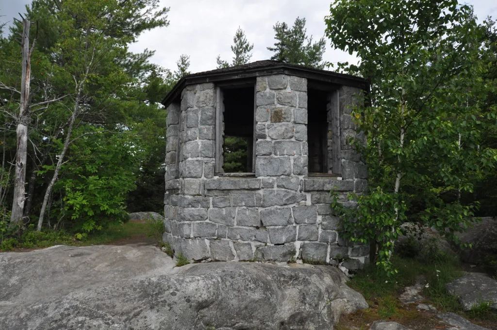 Small octagonal stone shelter with window openings on a rocky summit among birch trees