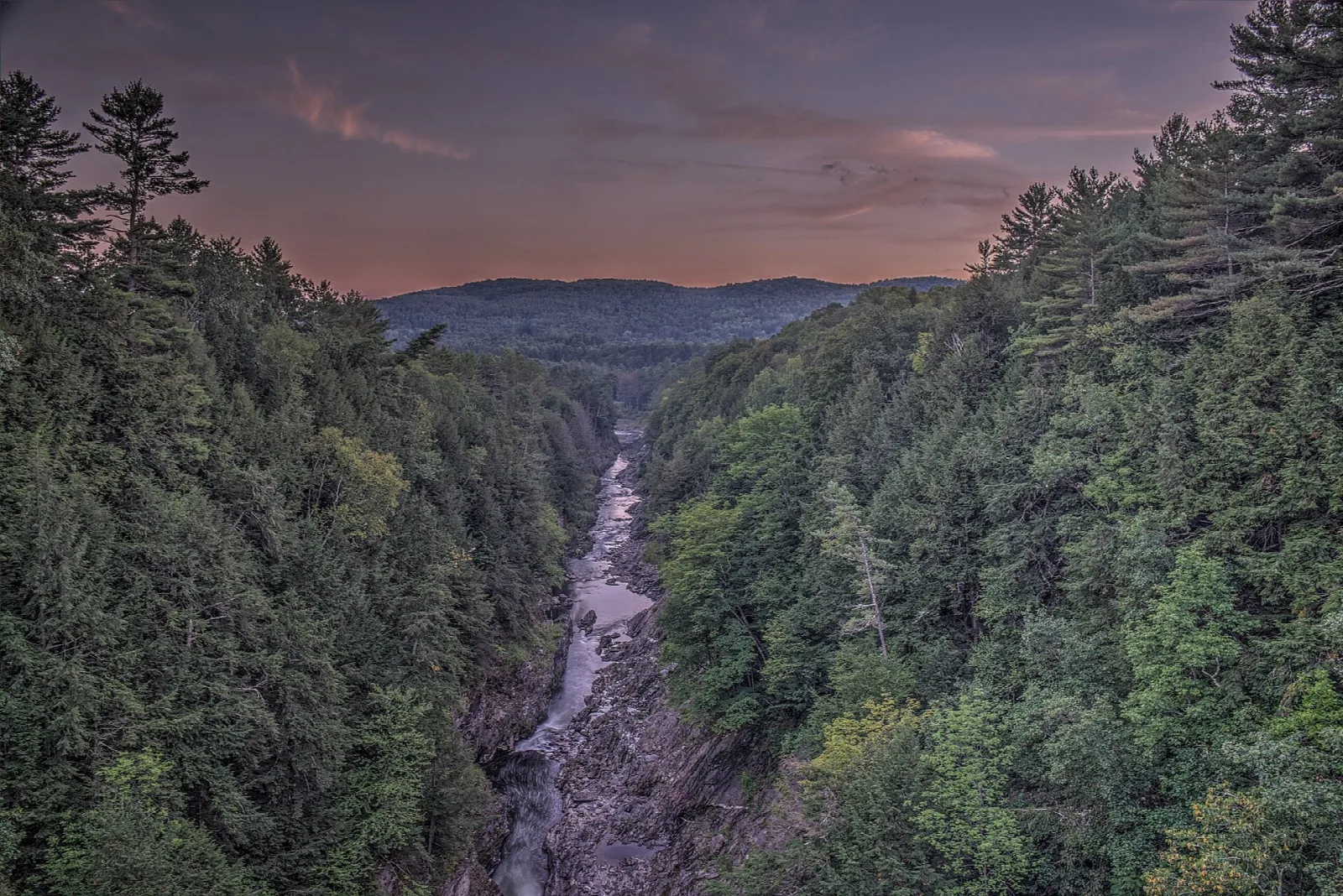Steep forested gorge at dusk with a river in the bottom and hills beyond