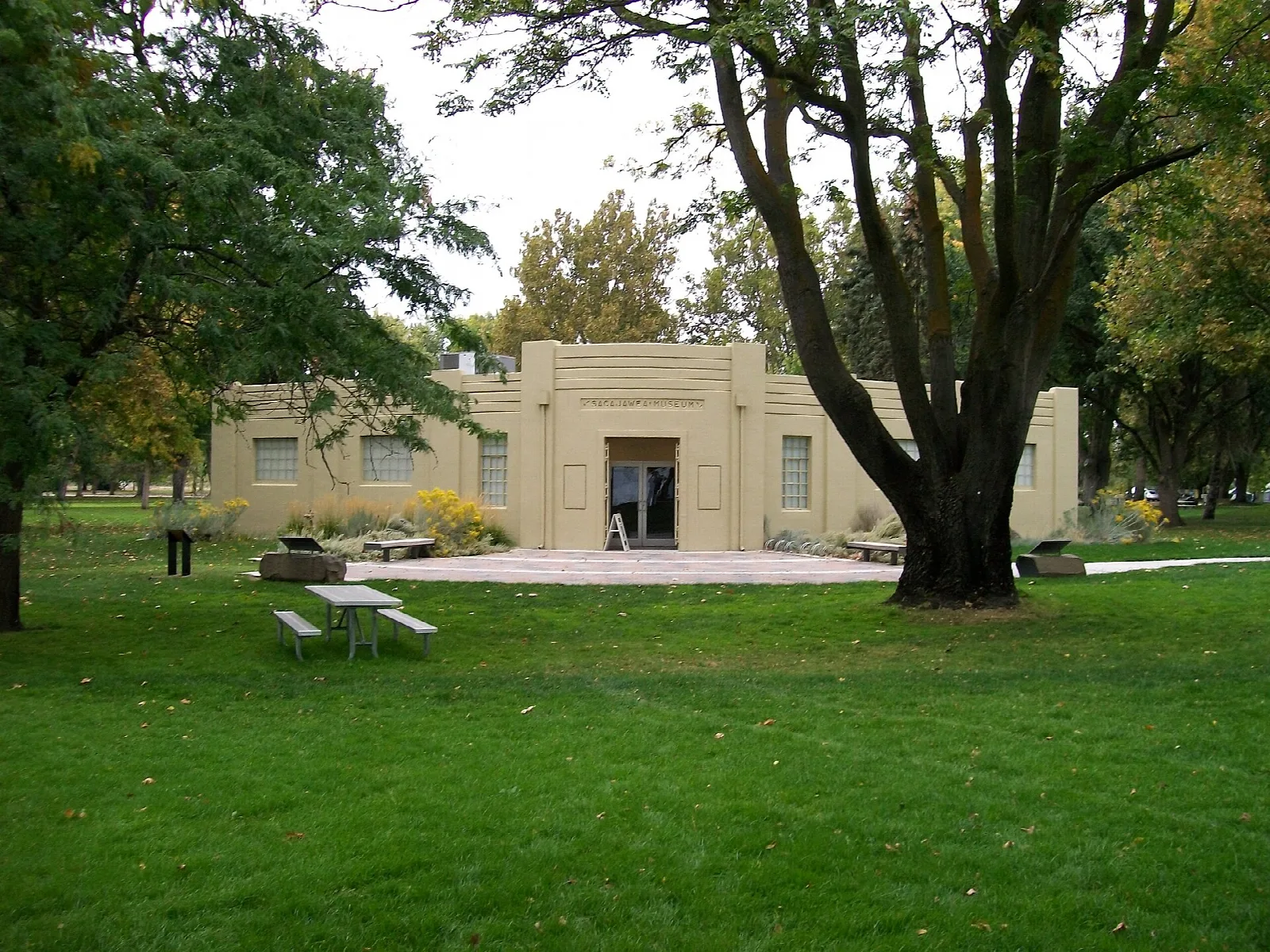 Low tan museum building behind a large tree and green lawn with picnic tables