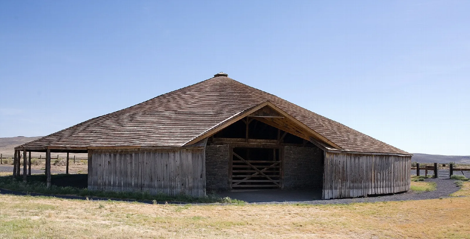 Round barn with a low shingled roof on open grassland under blue sky