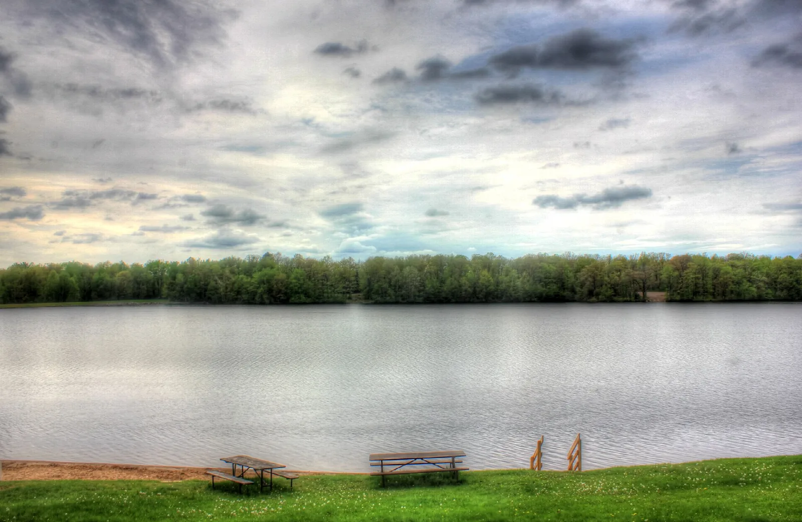 Lake with picnic tables on a grassy shore under a cloudy sky