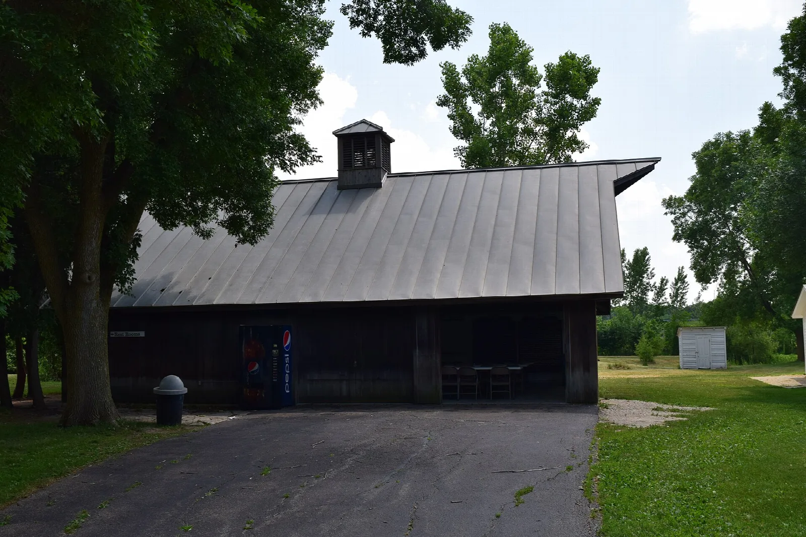 Long dark wooden barn with a metal roof beside a paved lane and trees