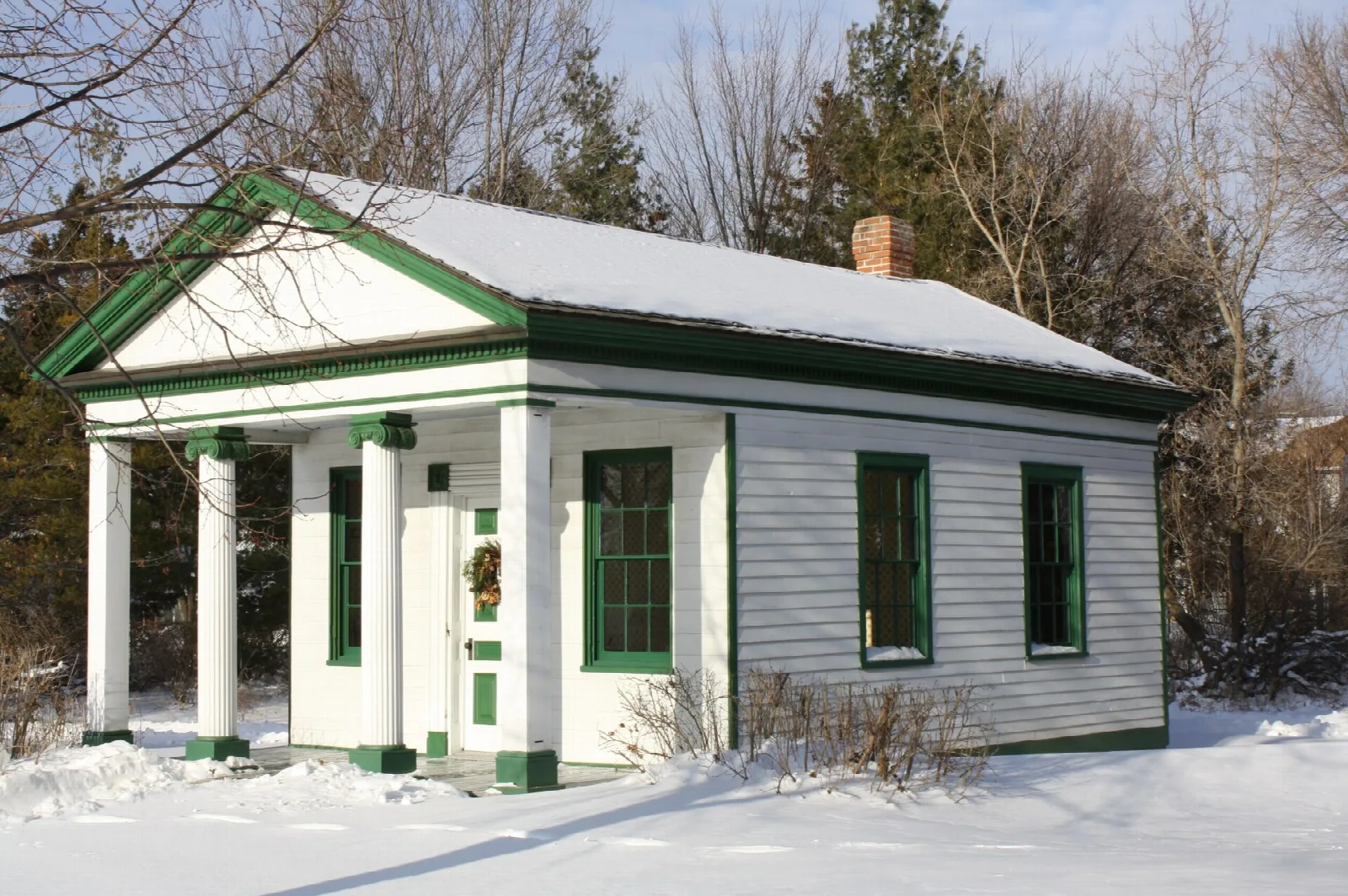 Small white building with columns and green trim, roofed with snow