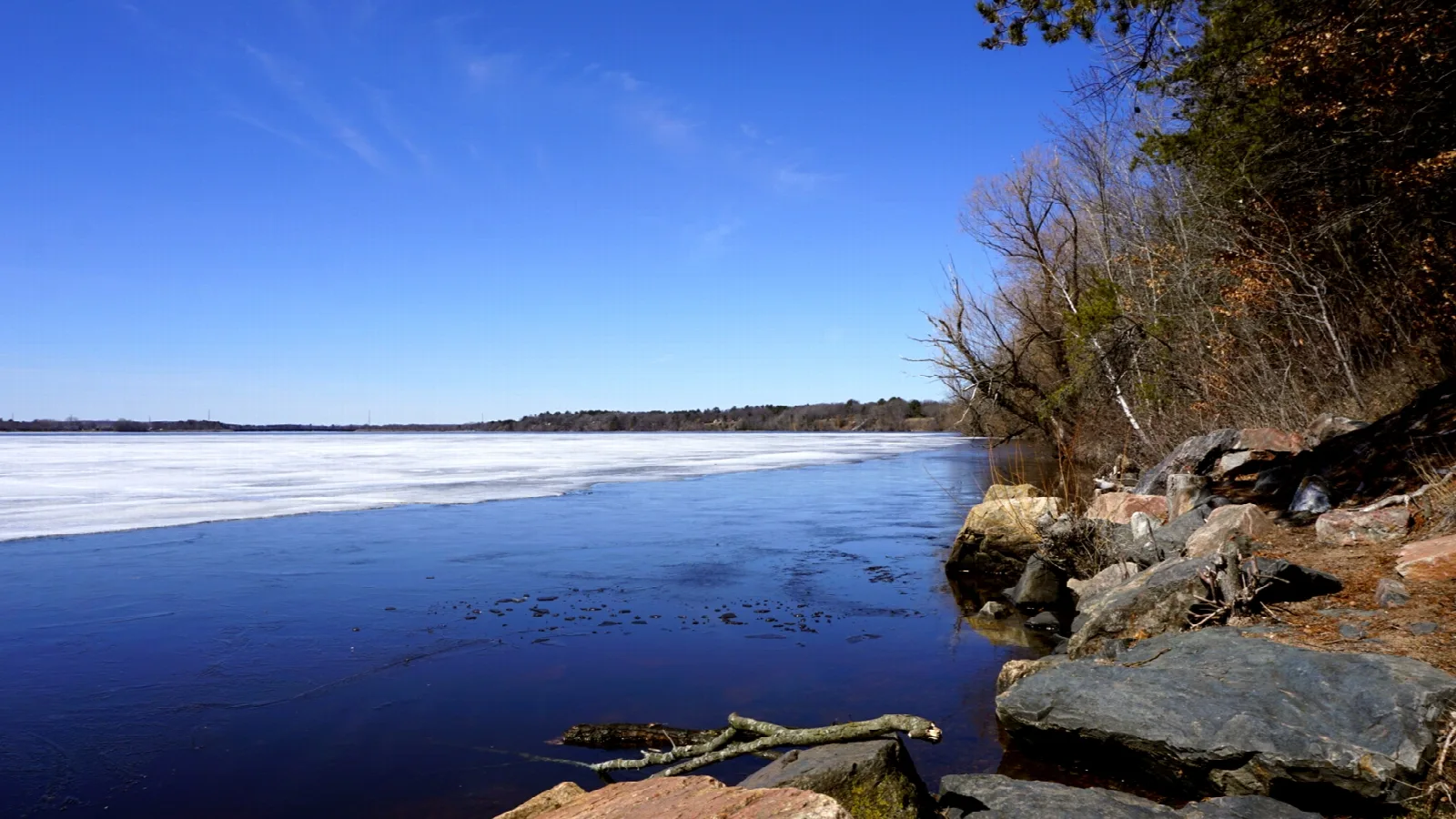 Blue lake with thawing ice and a rocky wooded shoreline under clear sky