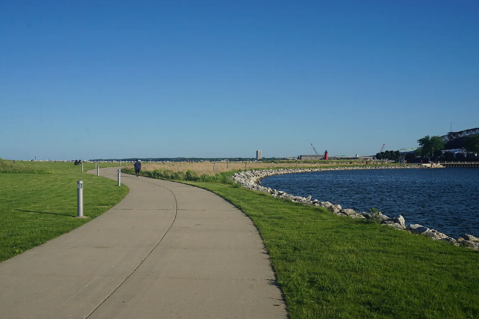 Paved path along grass beside a calm harbor under clear blue sky