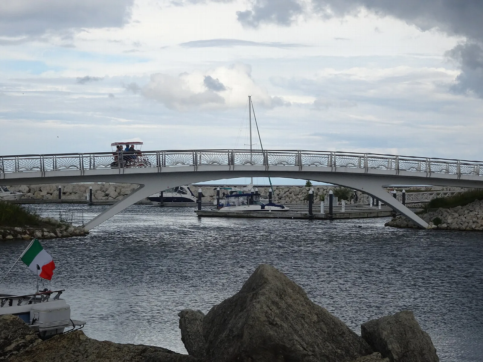 White arched footbridge over water with sailboat masts and large rocks in front