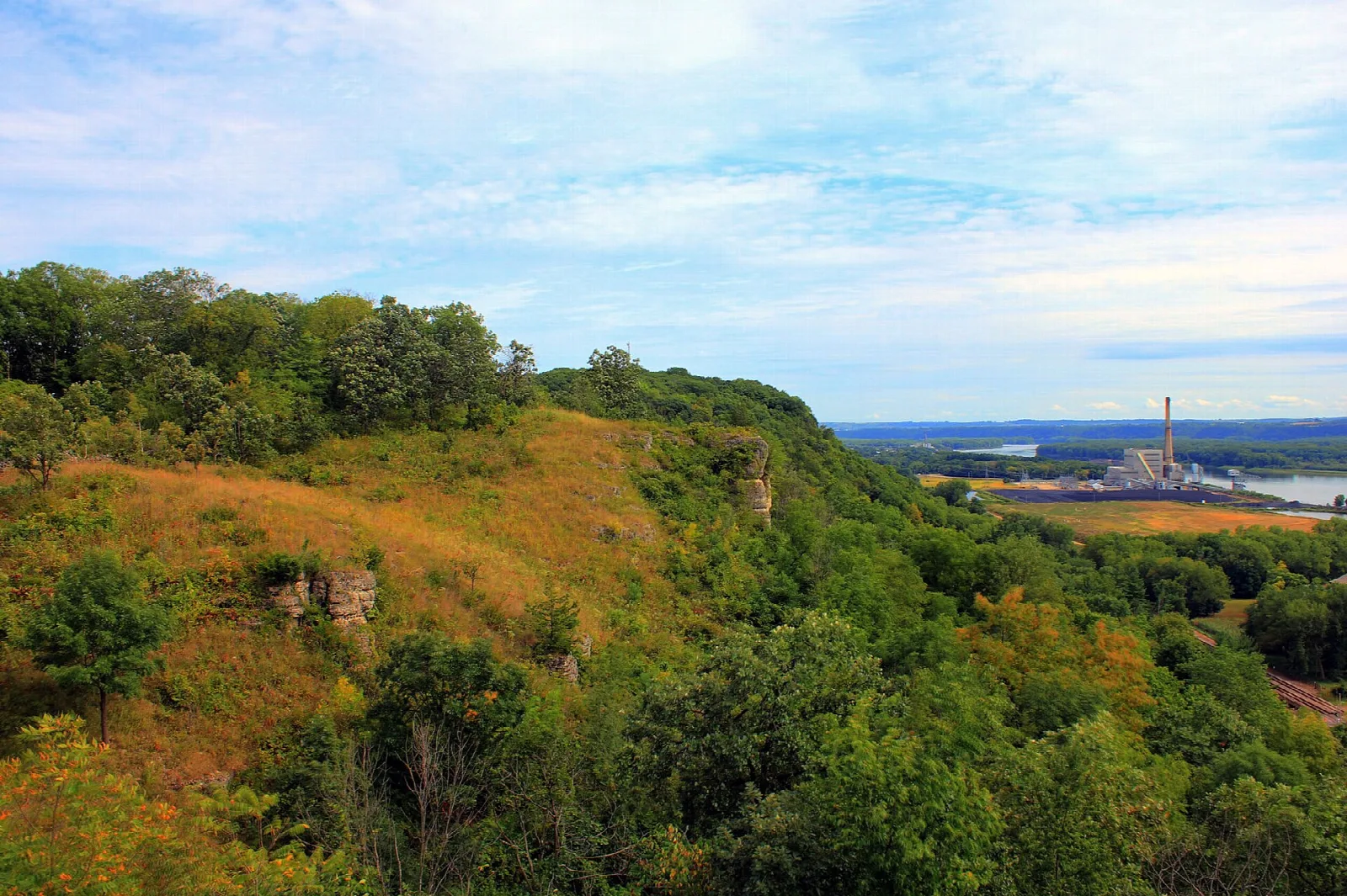 Grassy and wooded bluff above a river with an industrial plant in the distance