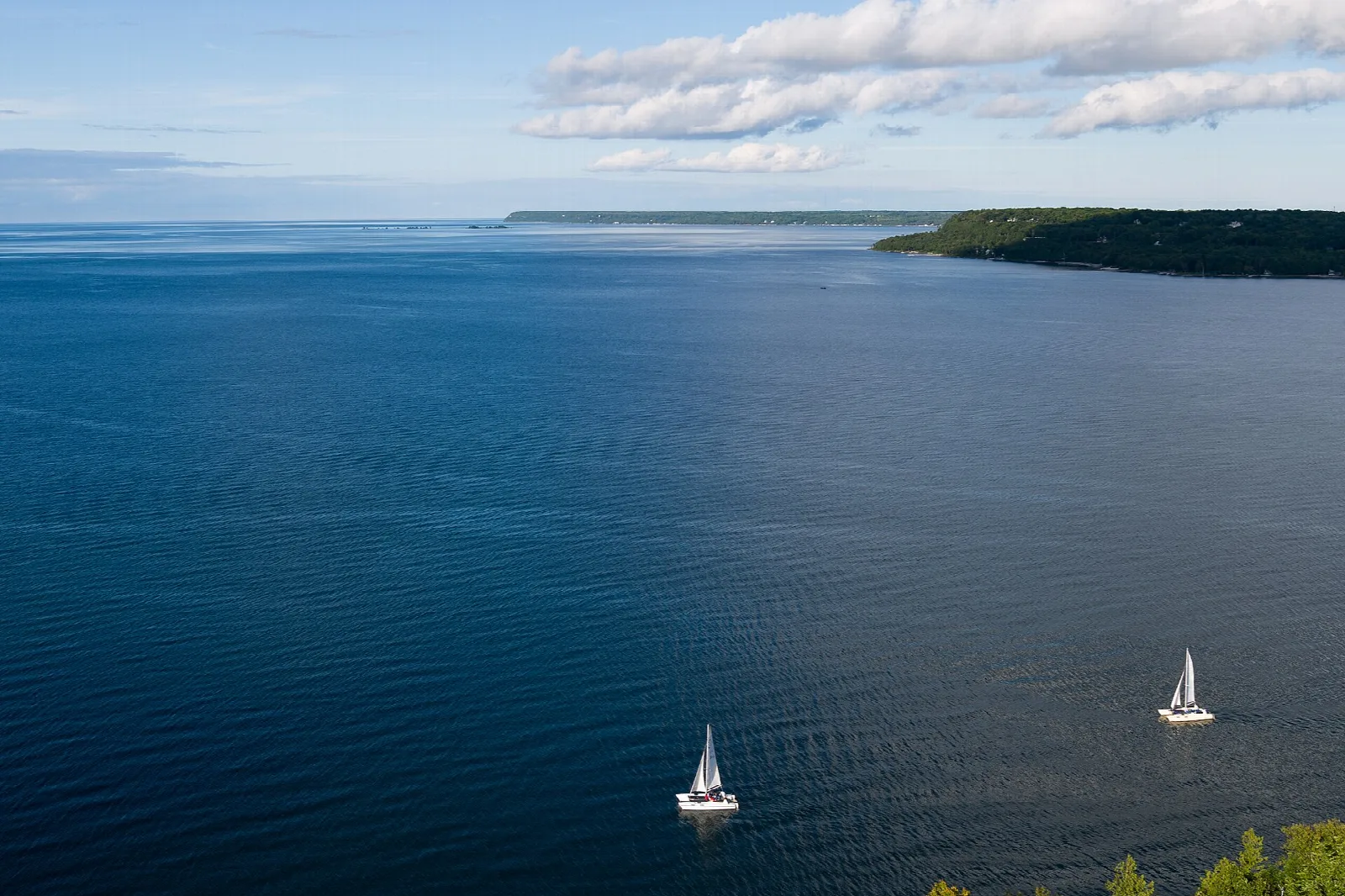 Wide blue bay with two sailboats and a wooded shoreline in the distance