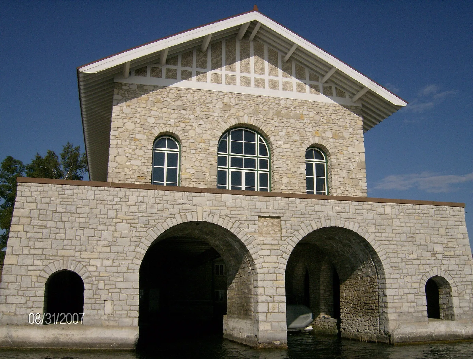 Stone boathouse with a gabled roof and arched water entrances against a blue sky