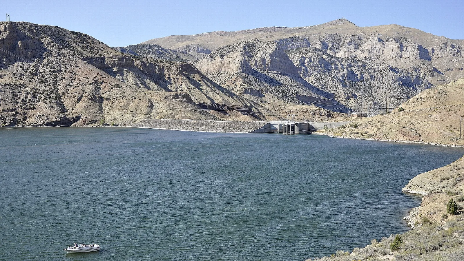 Blue reservoir with a small boat, dry brown hills and a dam in the distance