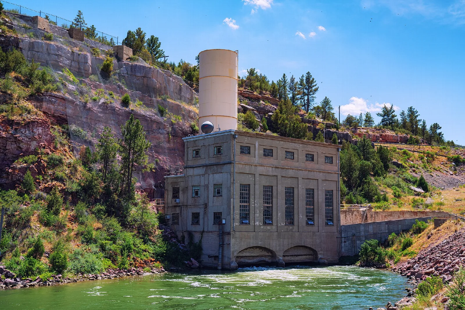 Stone power plant beside a green river below a rocky cliff
