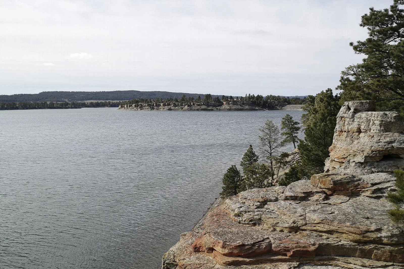 Gray lake seen from a sandstone ledge with pines, forested shore beyond
