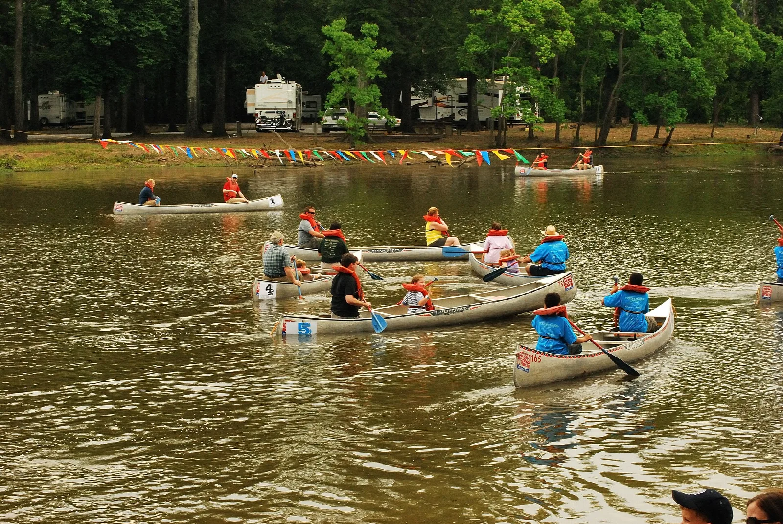 Several people paddling canoes on a lake, with trees and campers on the far shore