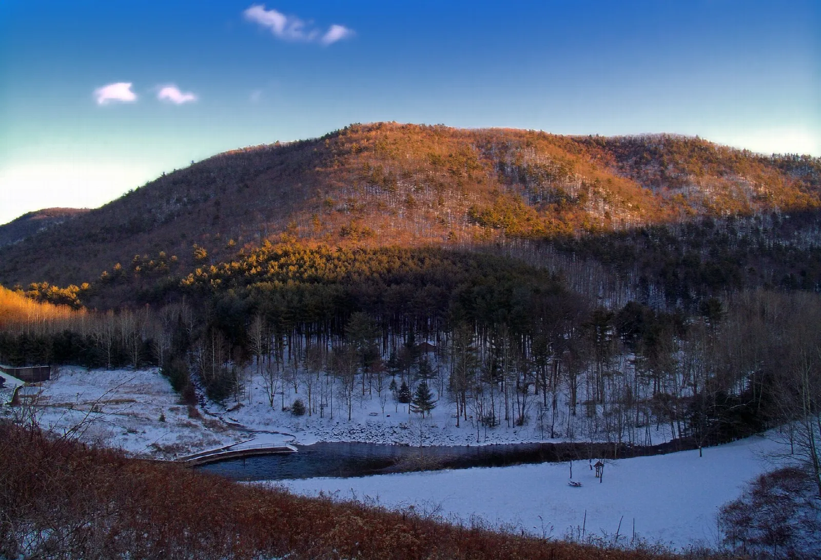 Forested mountainside lit orange at sunset with a snowy lakeshore
