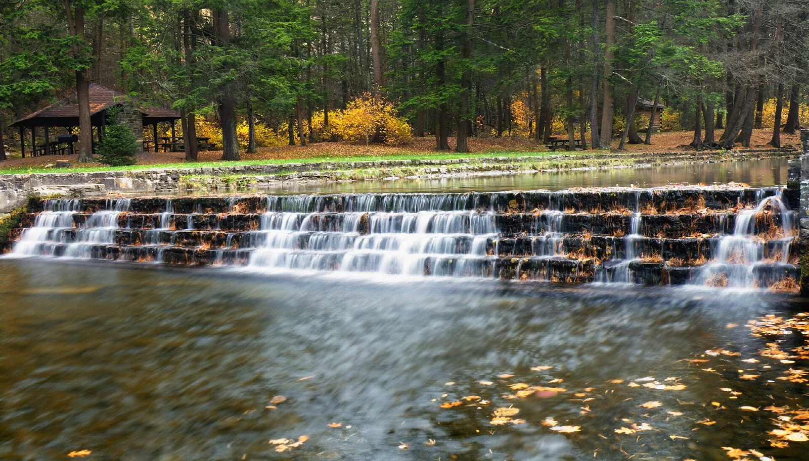 Low stone dam with water stepping over it beside a lawn and pavilion