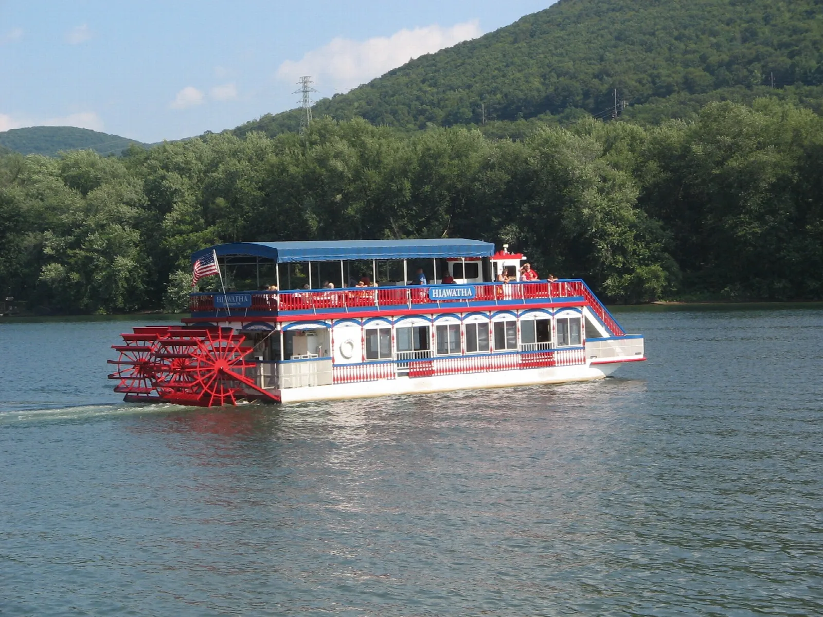 Red and white paddlewheel riverboat on a wide river below forested hills