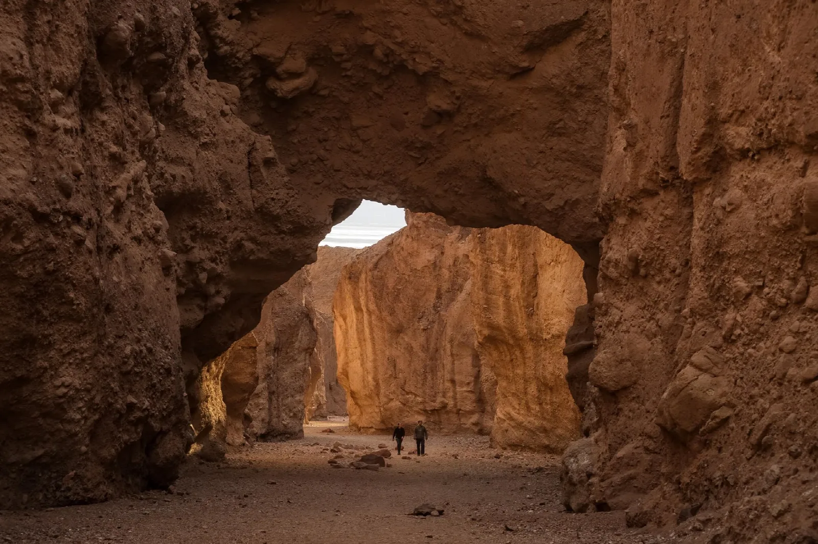 A massive rock span bridging a narrow desert canyon, with two hikers beneath