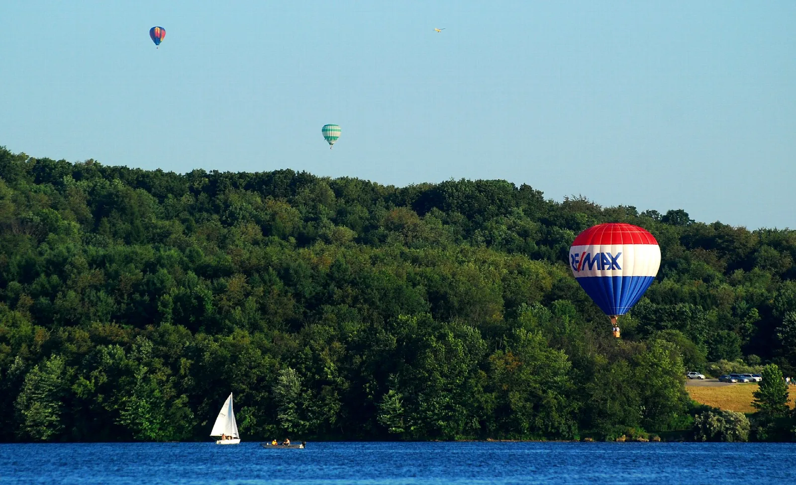 Hot air balloons and a sailboat over a blue lake with forested shore